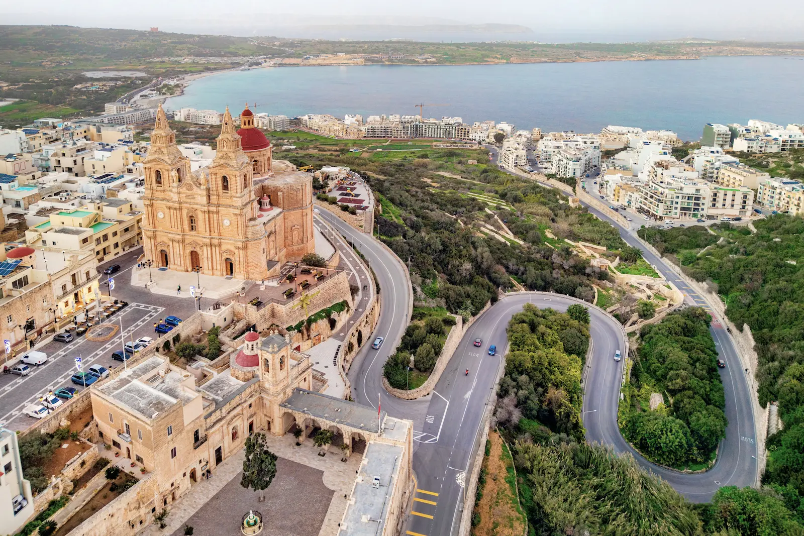 Luftaufnahme von Mellieħa auf Malta mit der Pfarrkirche im Vordergrund, der steilen Serpentinenstraße und der weitläufigen Mellieħa Bay im Hintergrund.